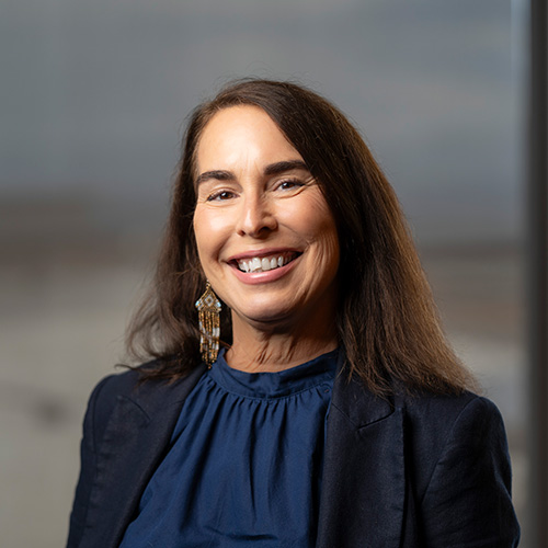 Wanda smiling at the camera. She is wearing a black blazer and blue blouse with a blurred background inside a building